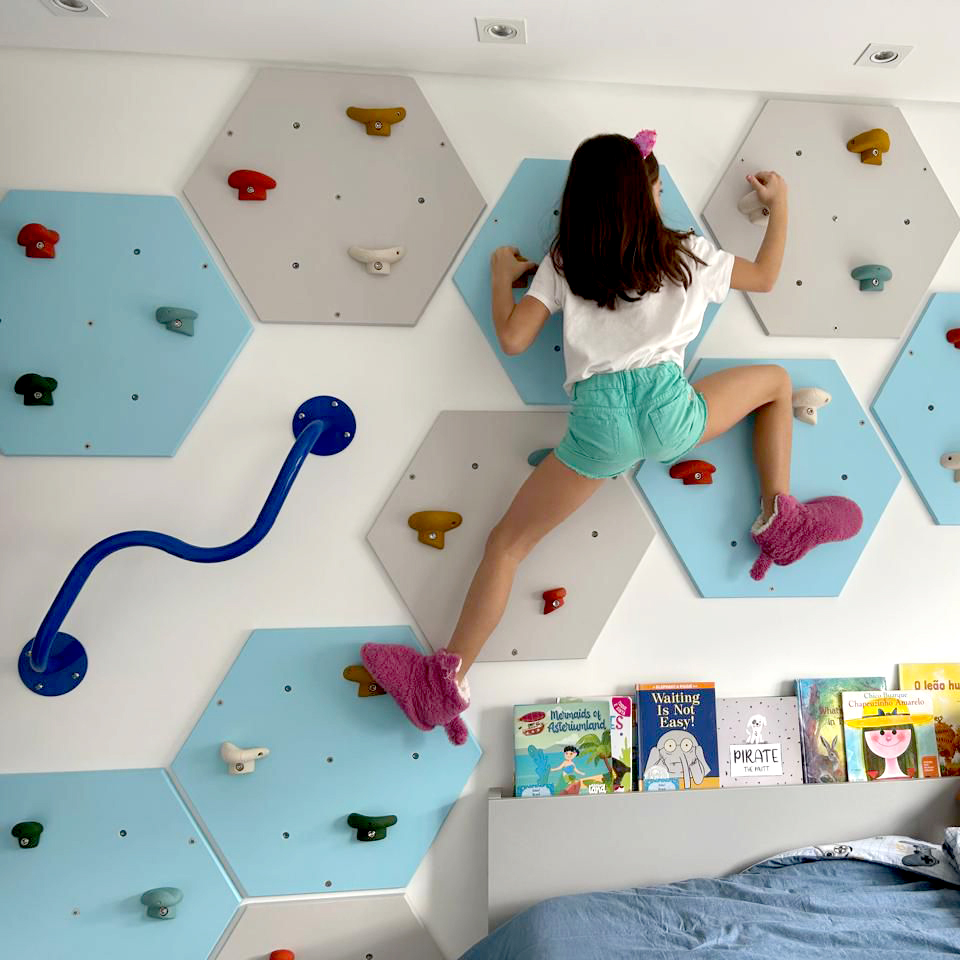 Parede composta por painéis de escalada e algumas barras metálicas enquanto uma criança brinca no quarto